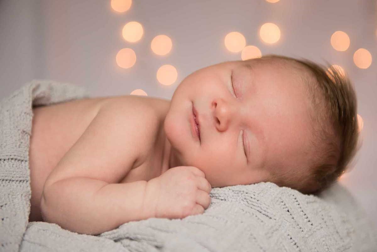 baby sleeping, wrapped in knitted blanket with twinkle lights in background